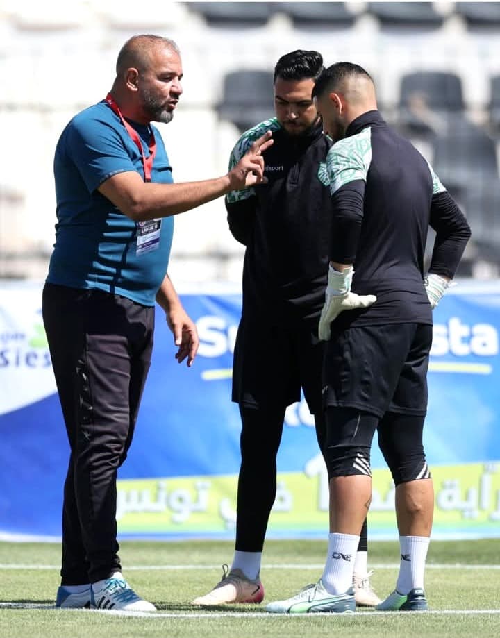 Raouf Rattouli donnant des consignes à deux gardiens de but pendant une séance d'entraînement, panneaux de sponsors du stade en arrière-plan.